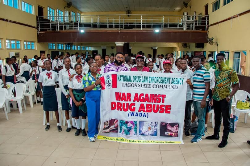 Students in school uniform and the Who I Go Be team in a school hall, standing around a National Drug Law Enforcement Agency, Lagos State Command banner that reads War Against Drug Abuse.